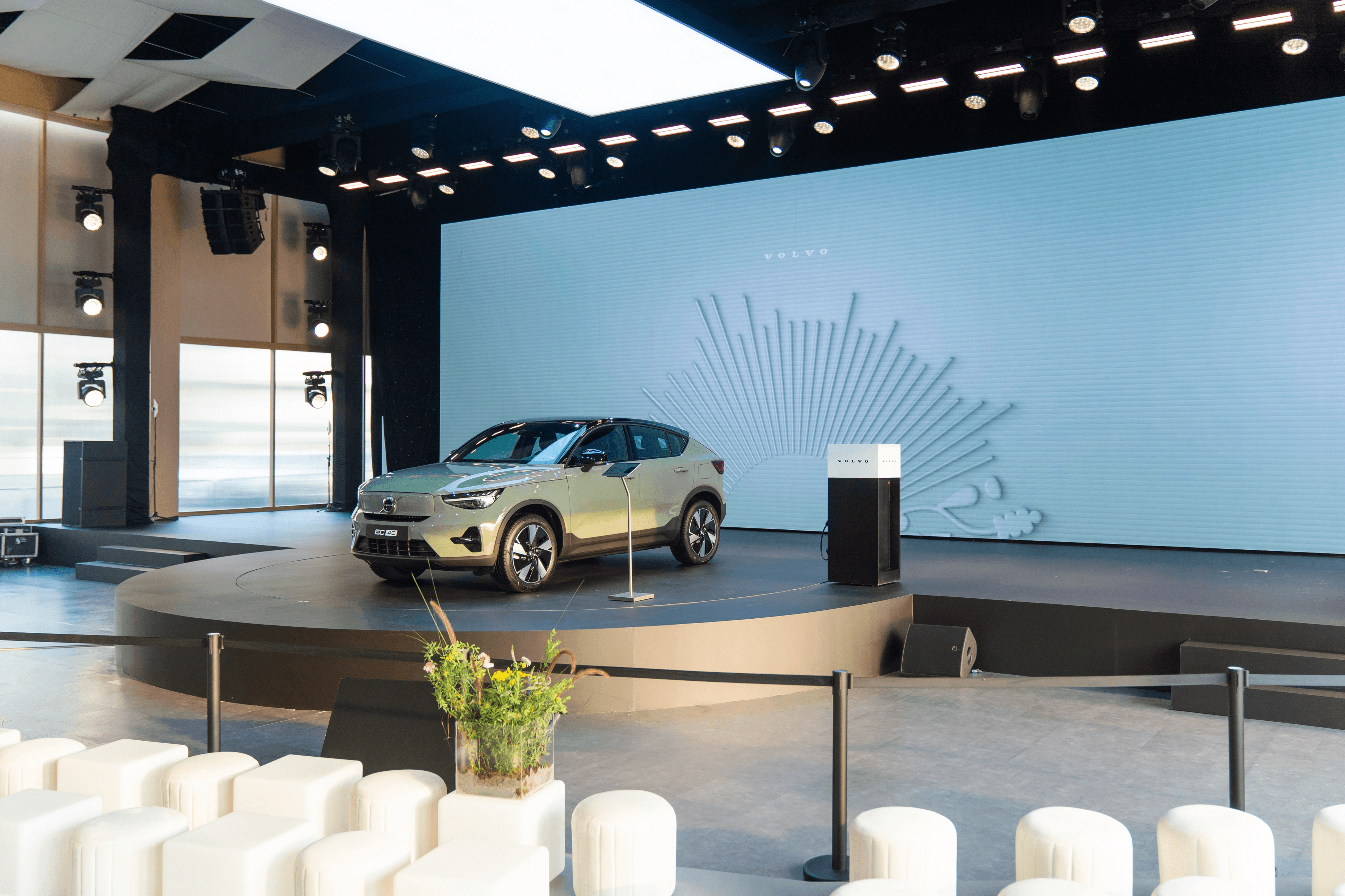 The event stage featuring the Volvo car parked next to a podium. The large screen behind it displays a clean, minimalist white line-art design alongside the Volvo logo.