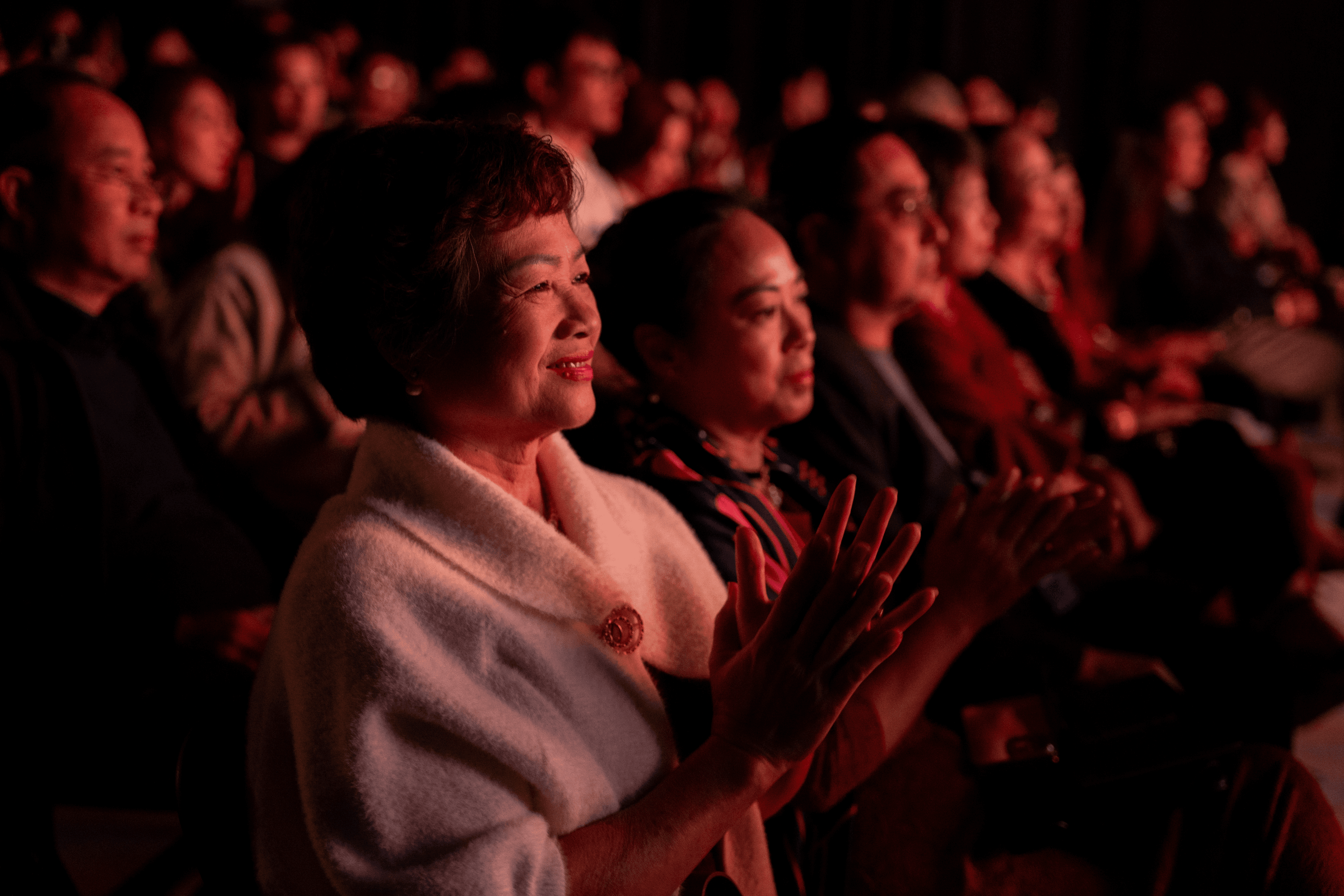 A shot of the audience bathed in a warm red light from the stage, focusing on a smiling woman in a white jacket enthusiastically clapping her hands.