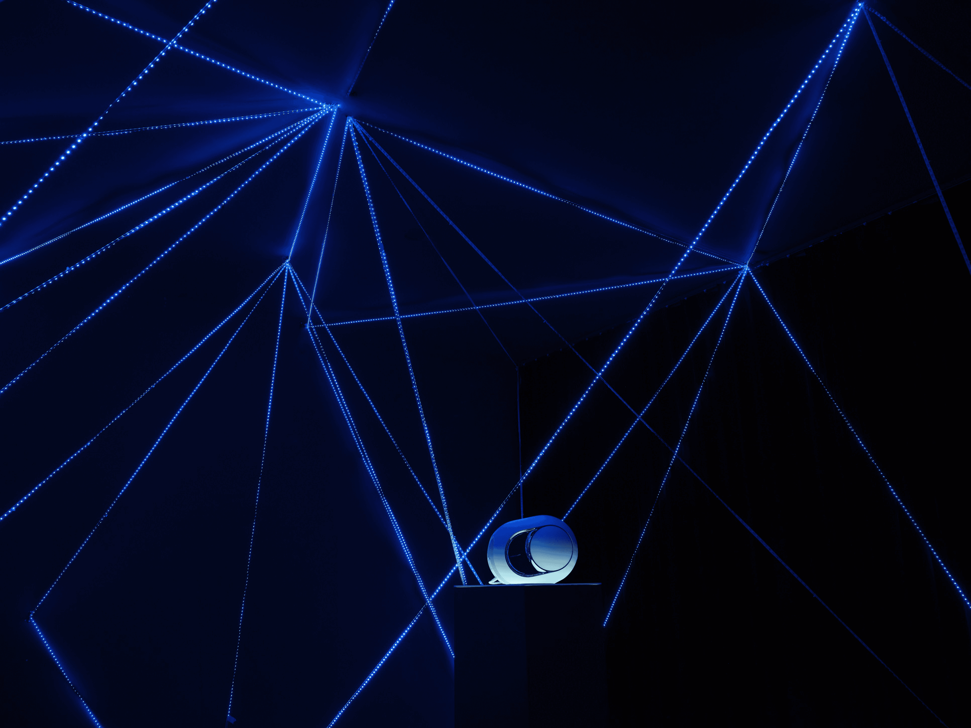 Close-up of the speaker source at the center of a geometric network of glowing LED sculpture radiating across the ceiling and walls of the exhibition