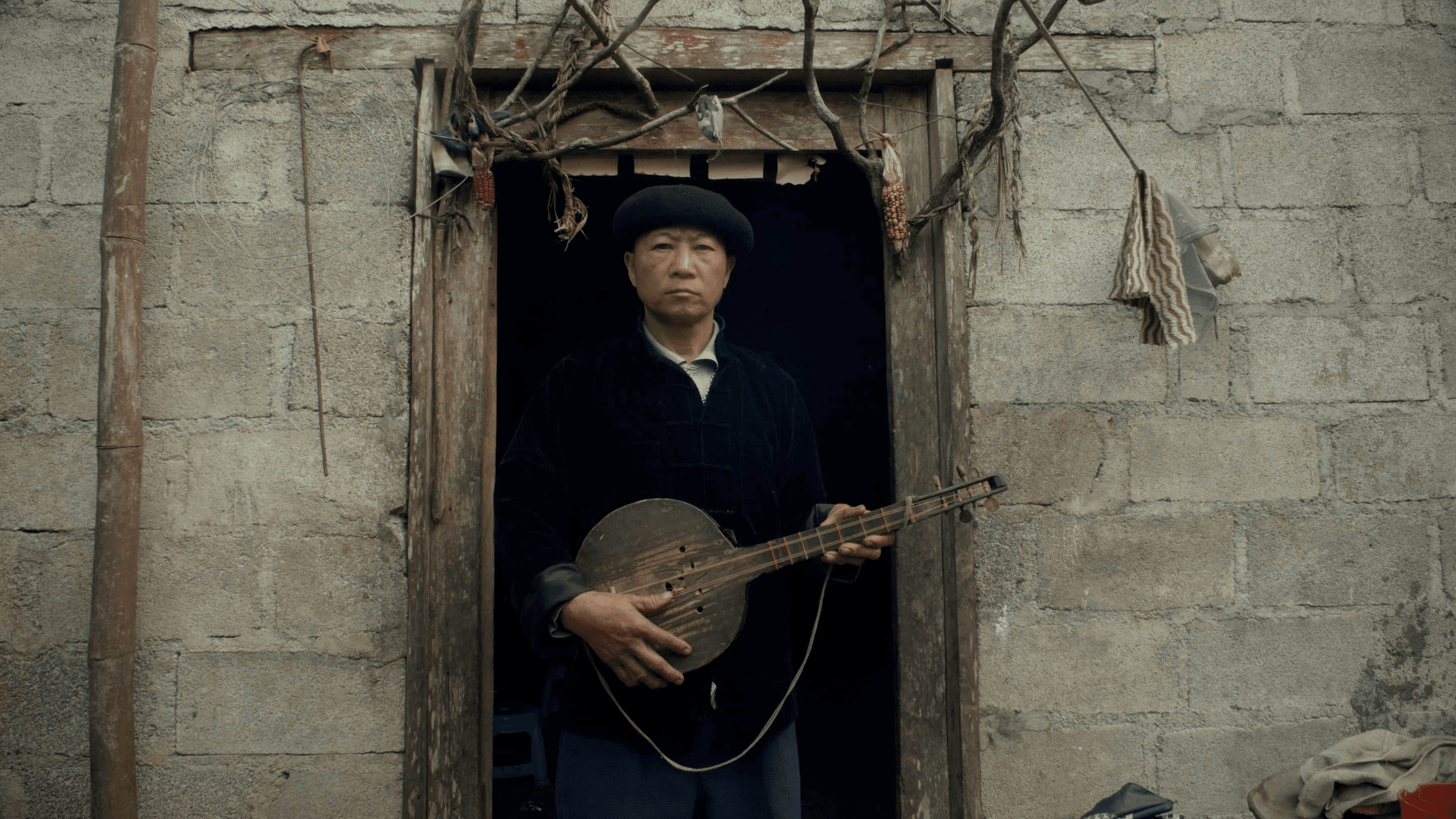 An older man in a black beret and dark jacket stands in a stone doorway holding a Vietnamese traditional string instrument. He looks directly into the camera.