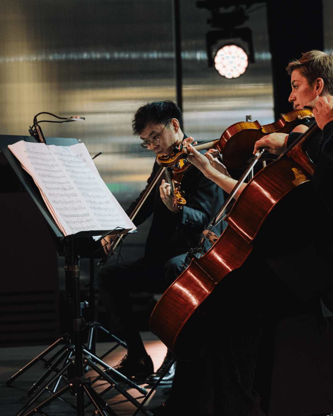 A close-up shot of the string quartet, focusing on the violinist Nguyễn Thiện Minh playing illuminated by a stand light, with a female violinist and a cello prominently in the foreground.