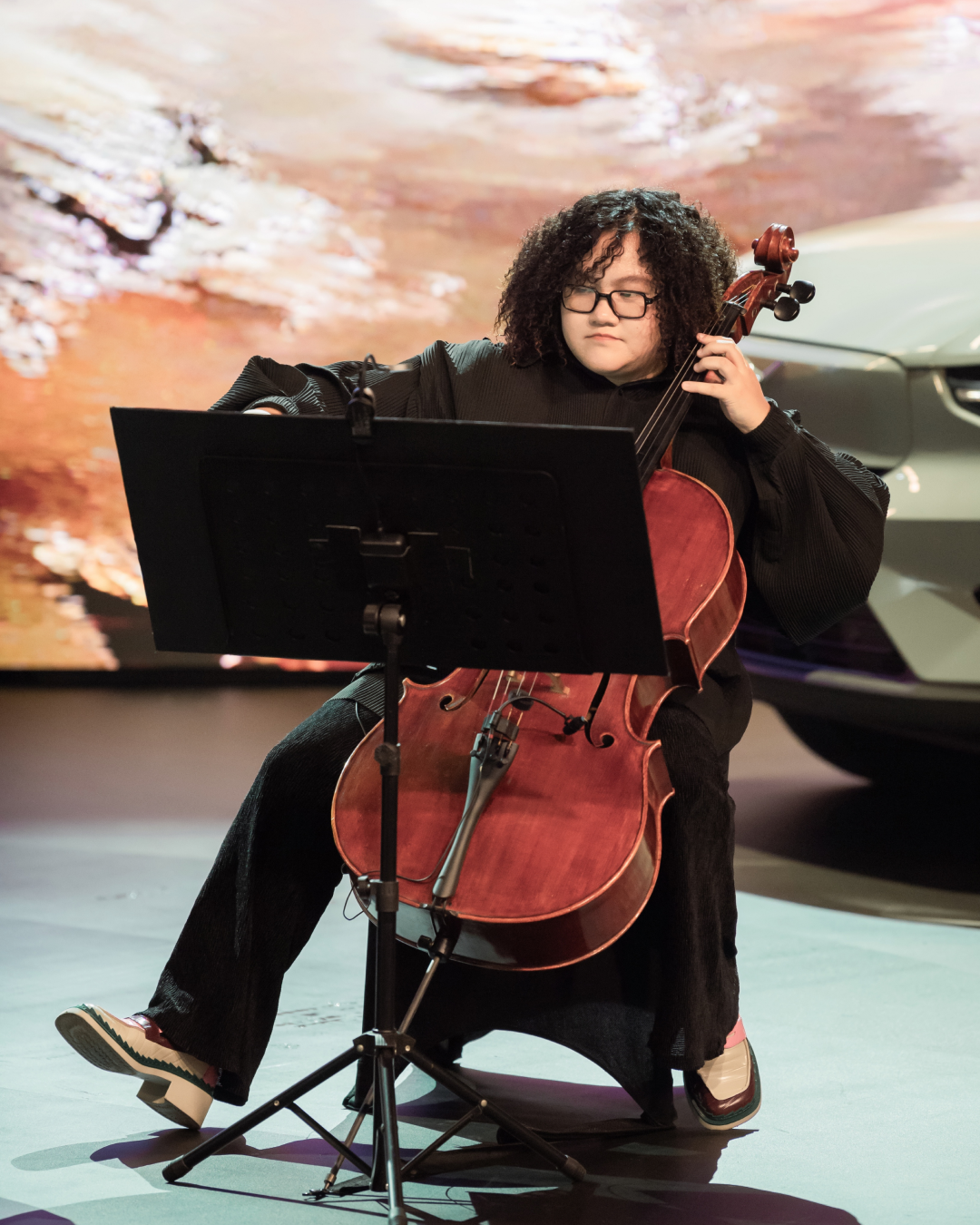 A focused shot of the cellist Trần Hồng Nhung performing on stage, with a portion of the green Volvo car and a warm, textured digital backdrop visible behind them.