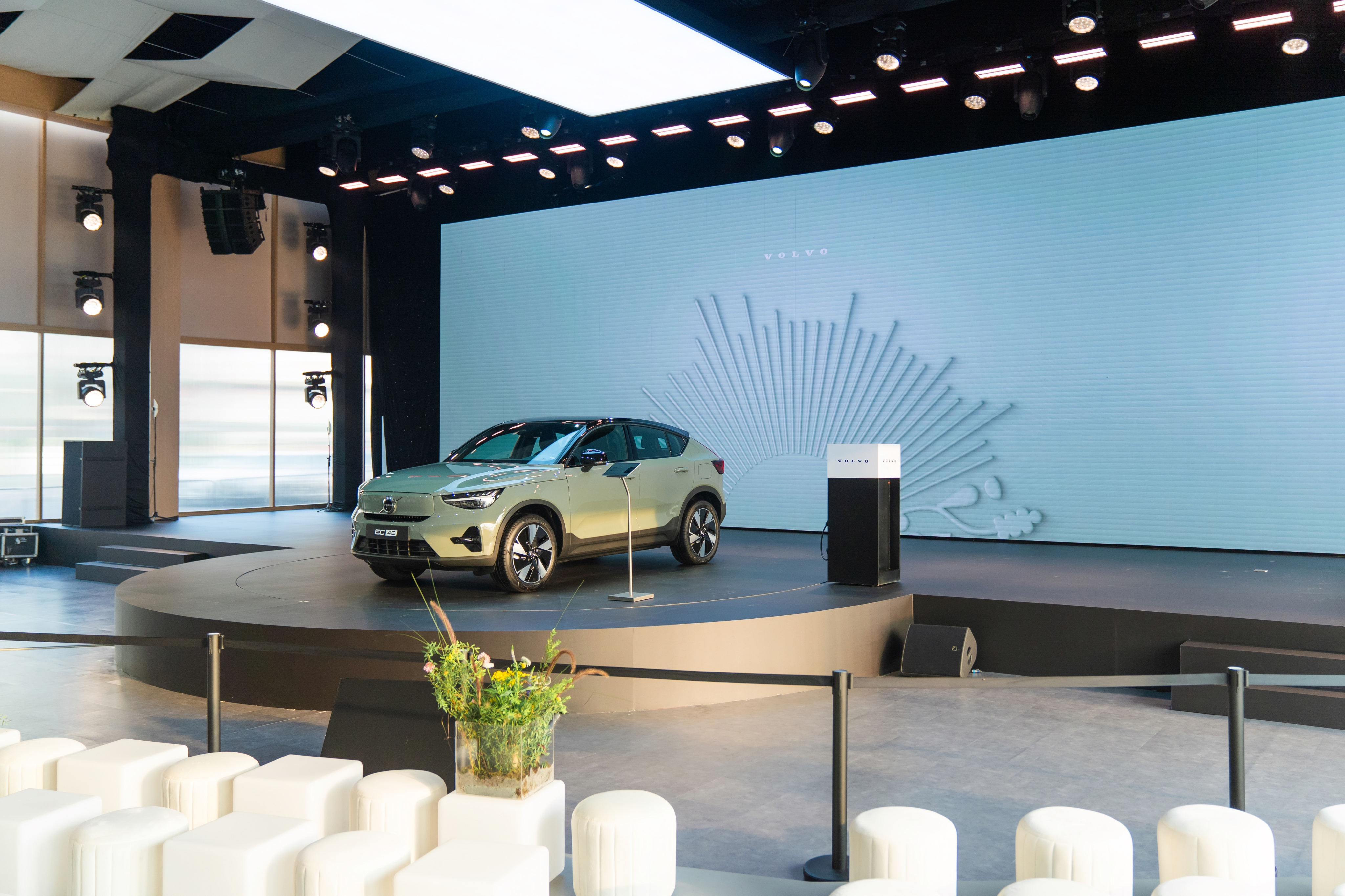 The event stage featuring the Volvo car parked next to a podium. The large screen behind it displays a clean, minimalist white line-art design alongside the Volvo logo.