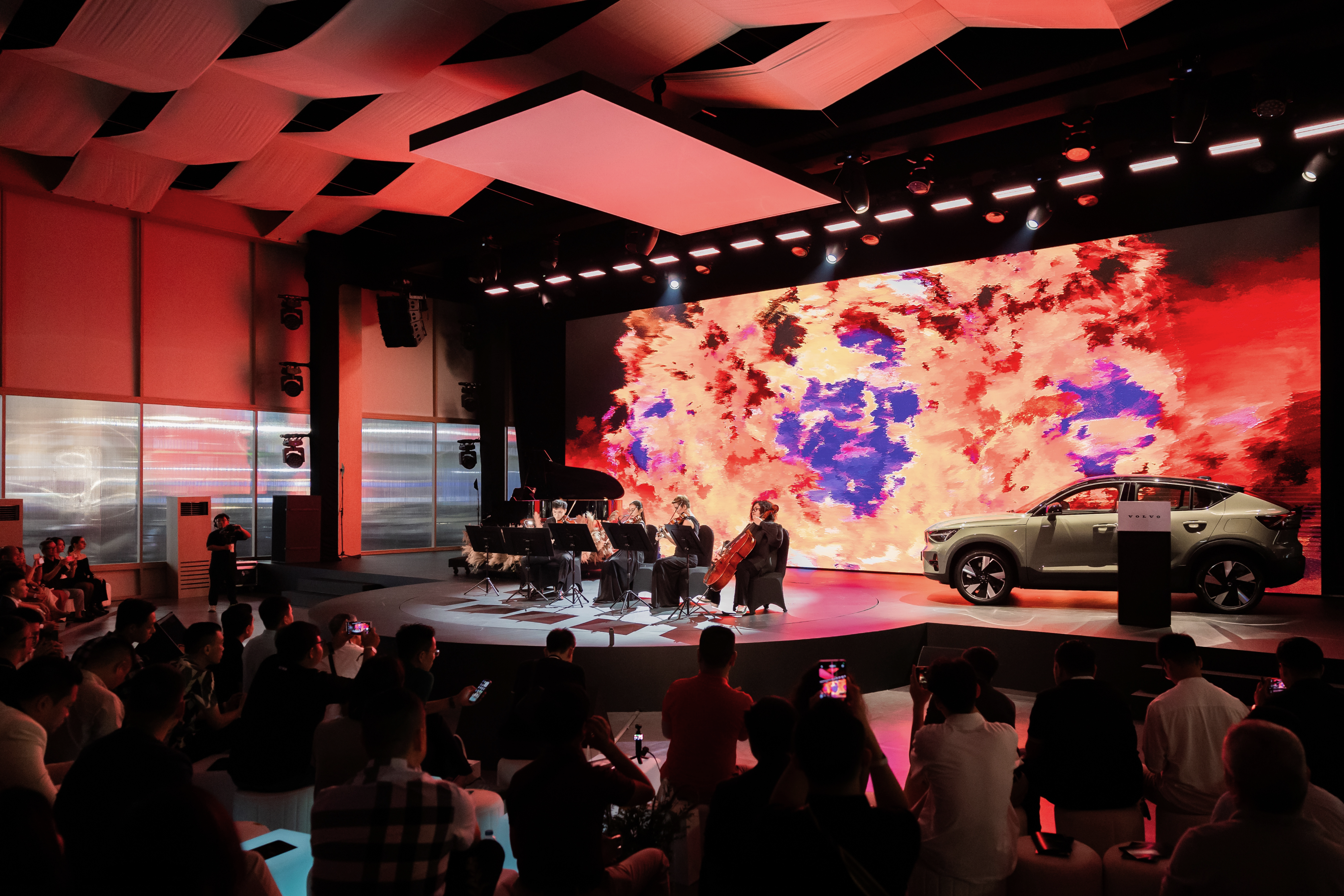 A wide shot of the event stage where a string quartet performs next to a Volvo car. The large LED background screen displays fiery red and orange generative art, with an audience watching from the foreground.
