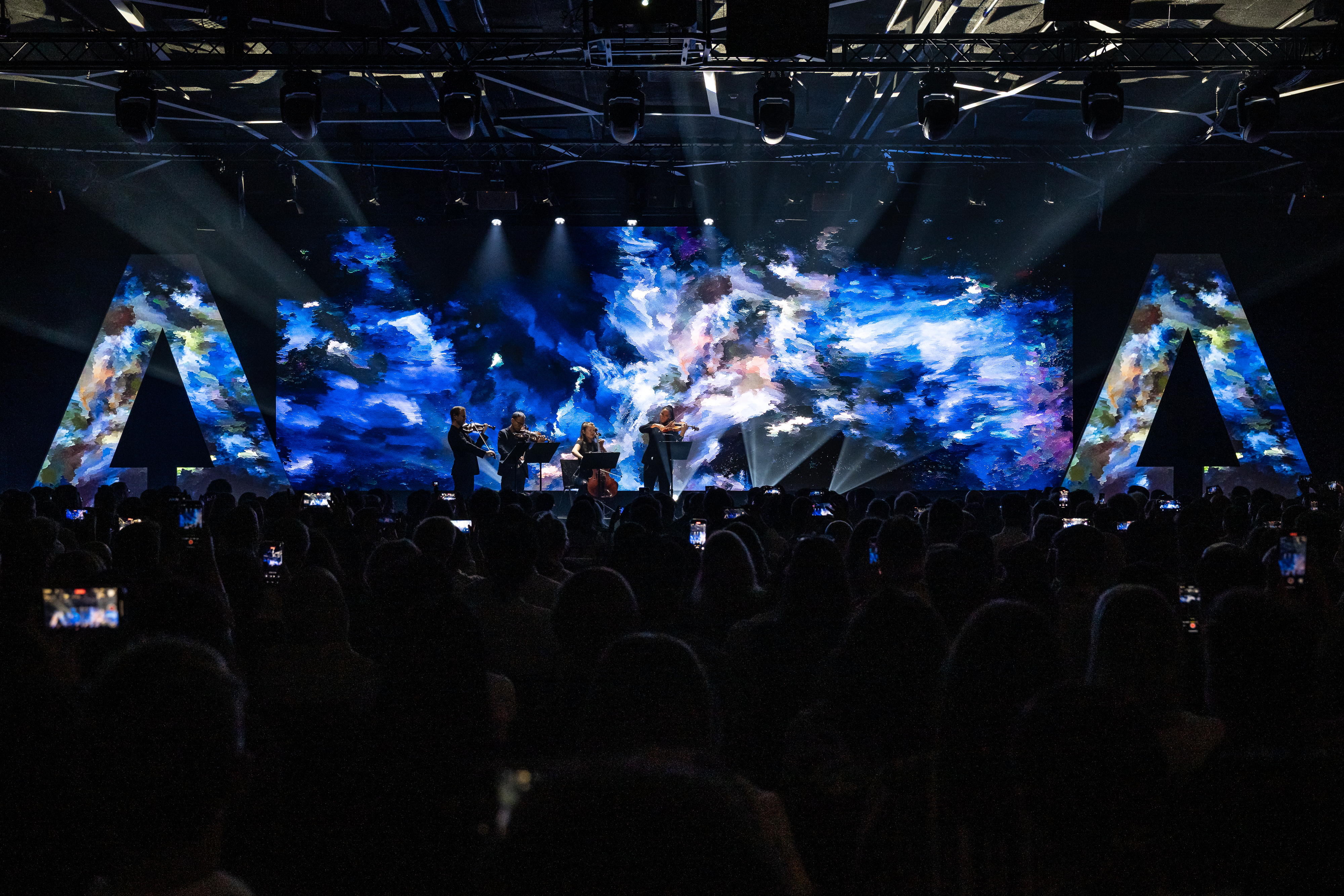 A large audience fills a darkened conference hall at the AI For Good Summit 2025, watching a string quartet perform on stage. Wide shot from behind the crowd.