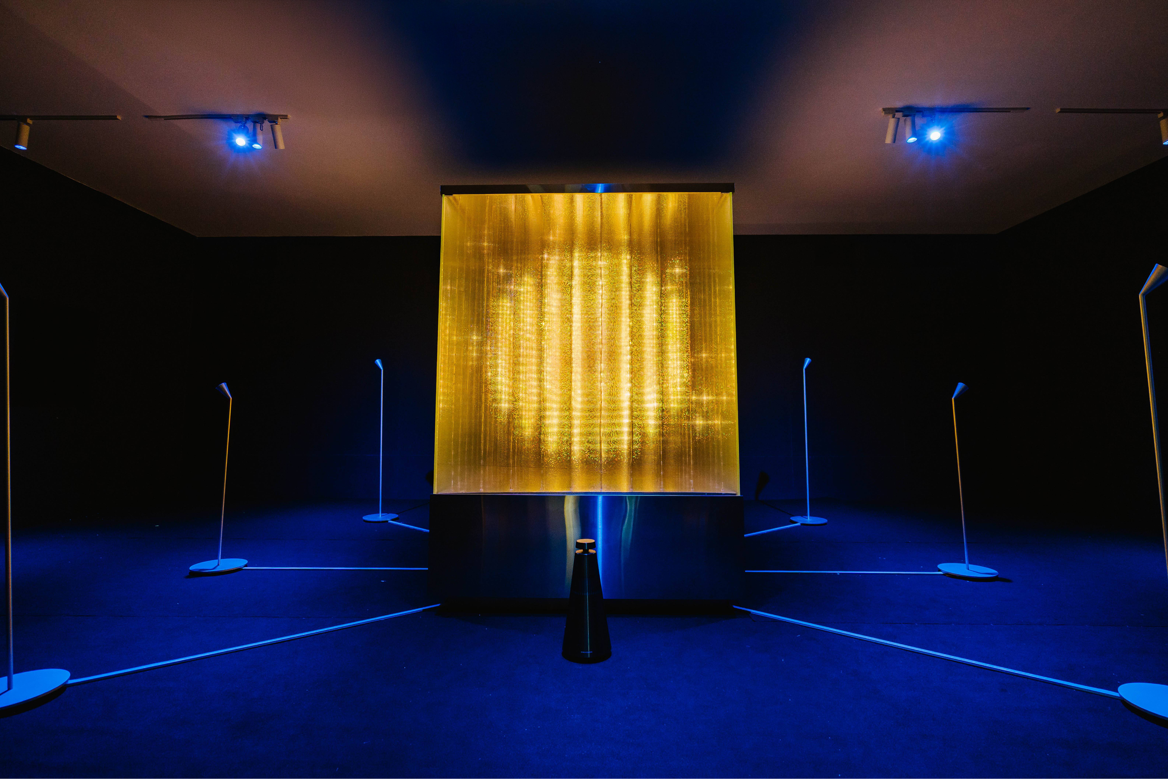 A dark exhibition room centered around a large, brightly glowing cube installation, with six tall microphone stands placed around it.