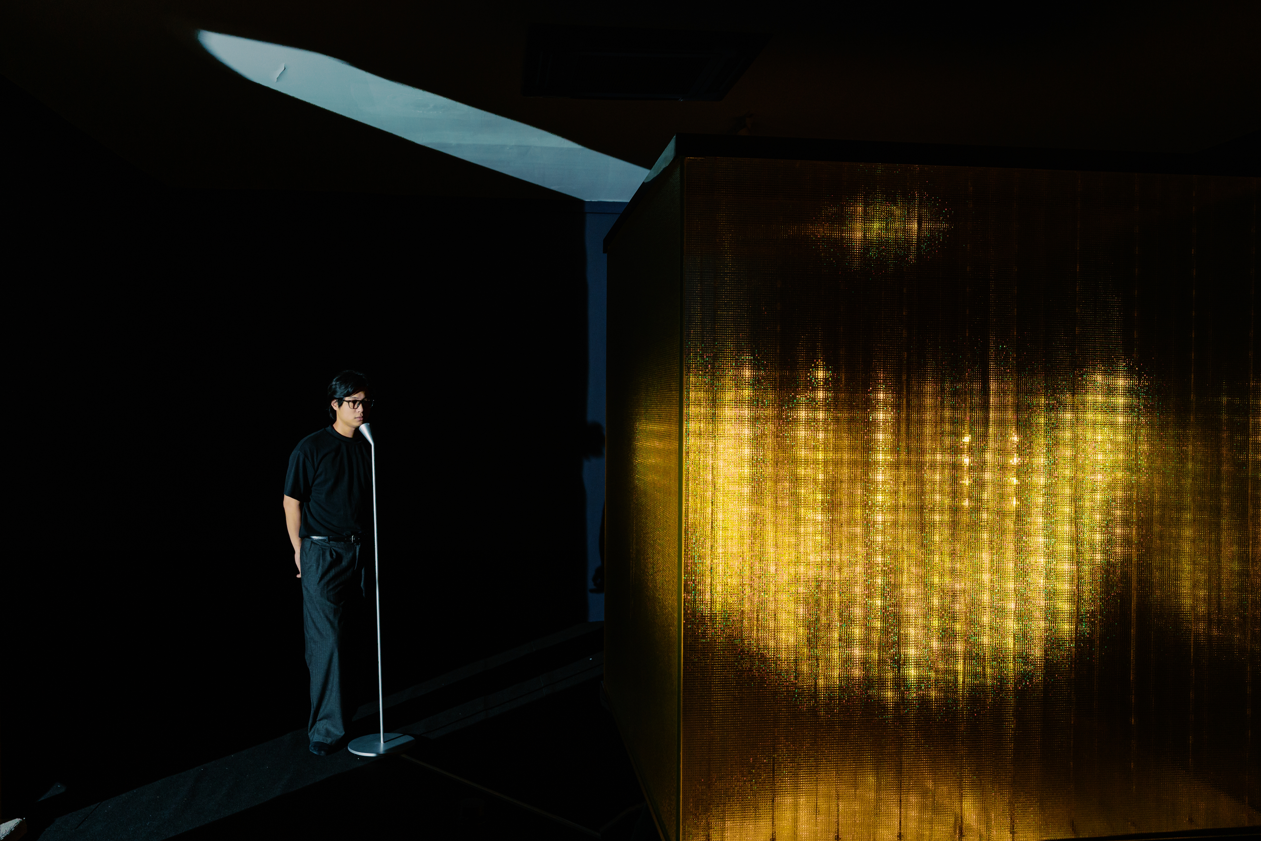 A person stands in a room, facing a white microphone stand positioned next to a large glowing cube installation.