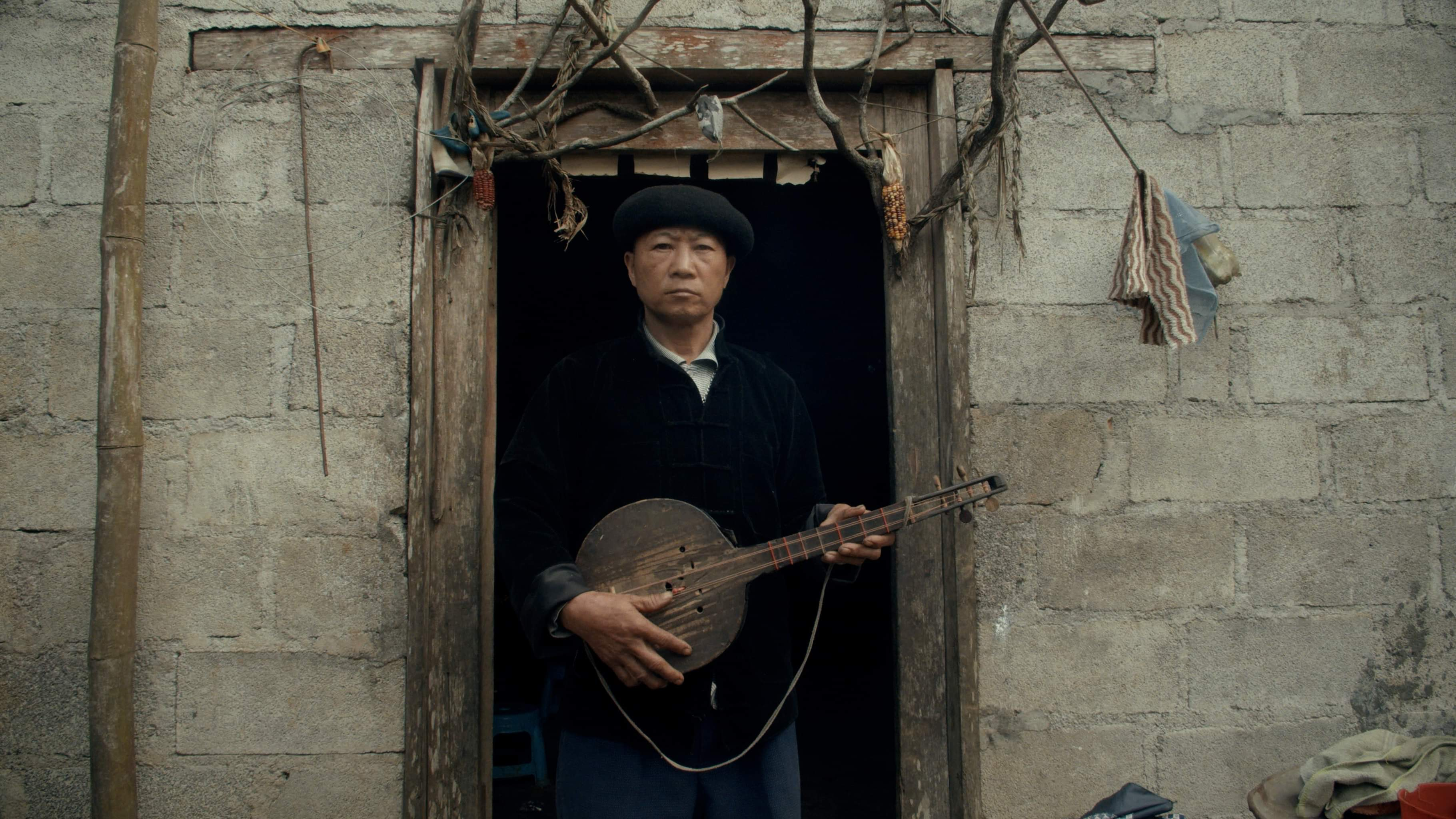 An older man in a black beret and dark jacket stands in a stone doorway holding a Vietnamese traditional string instrument. He looks directly into the camera.