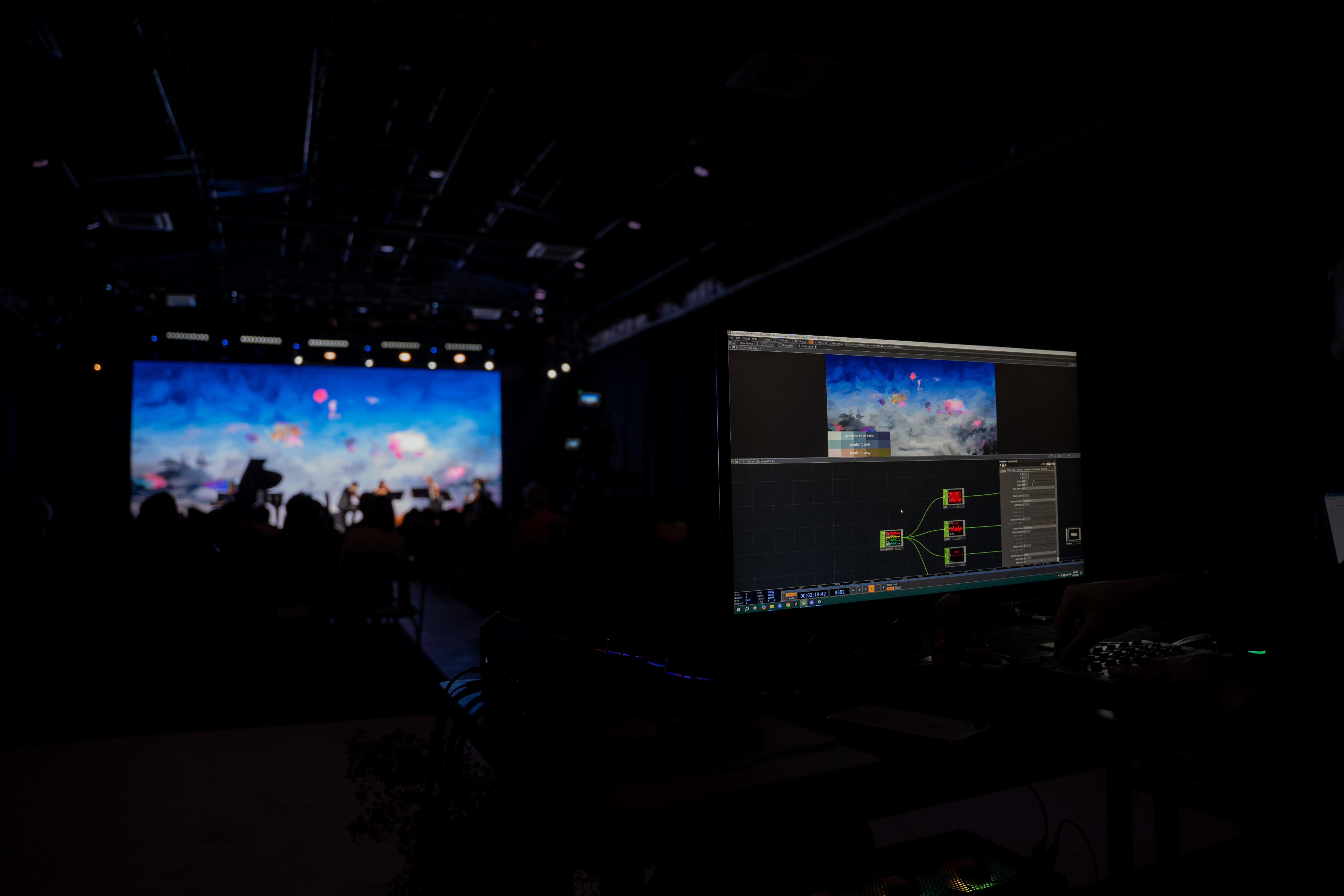 A behind-the-scenes view from the tech desk, showing node-based real-time system on a monitor controlling the generative visuals shifting on the distant main stage.