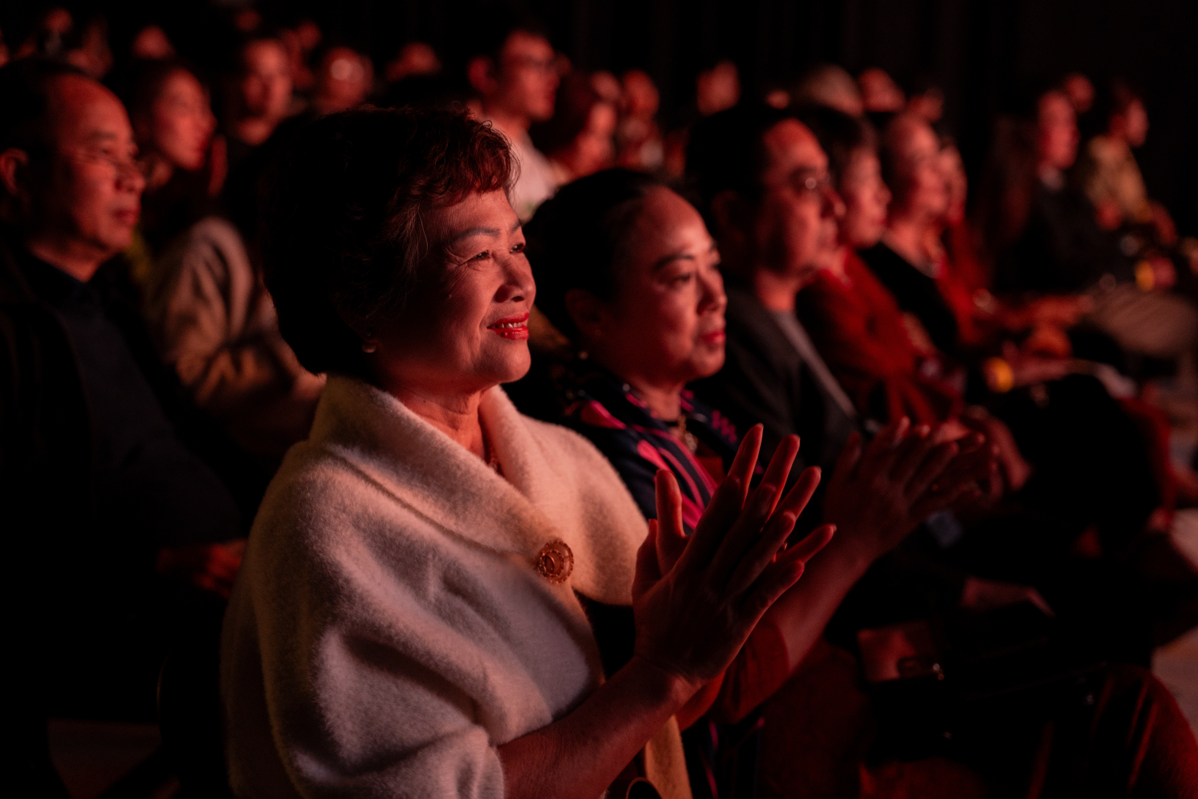 A shot of the audience bathed in a warm red light from the stage, focusing on a smiling woman in a white jacket enthusiastically clapping her hands.