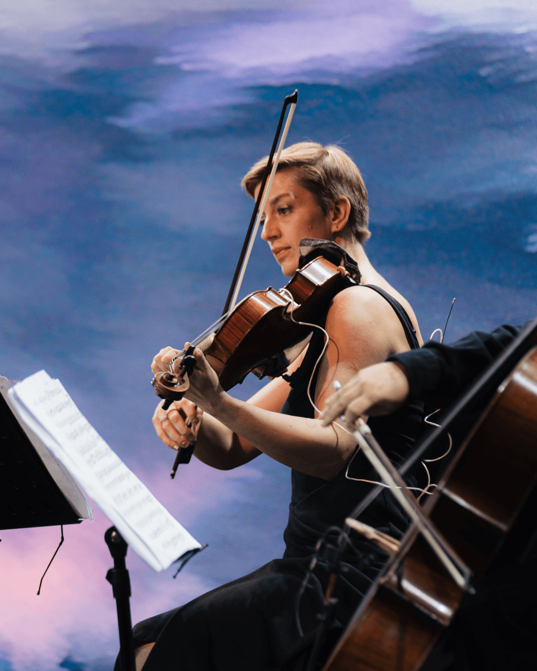 A close-up profile of the violinist Svetlana Golubovskaya playing with sheet music, illuminated by soft blue and pink generative art.