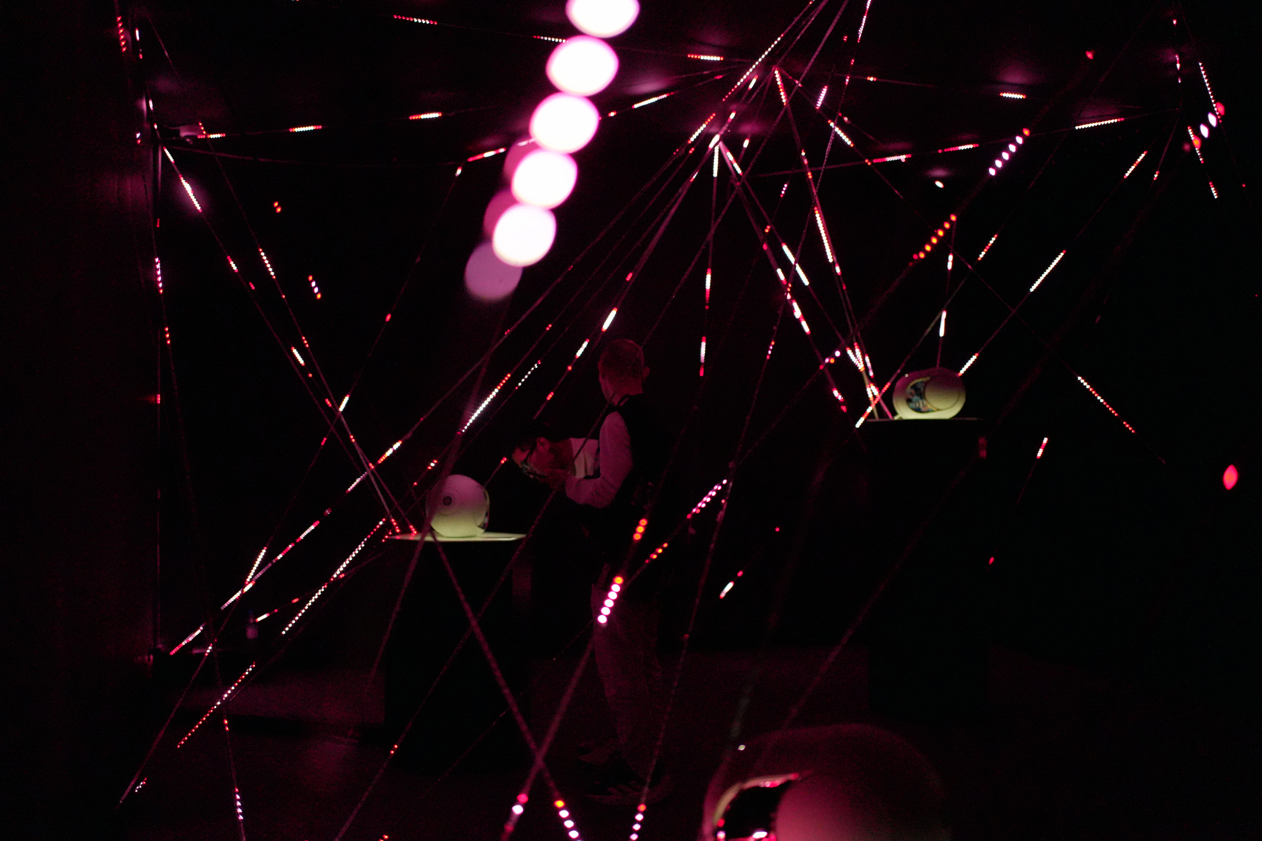 The visitors observe with a spatial sound source on a pedestal, surrounded by a web of red LED strings in the 'Silent Symphony' installation